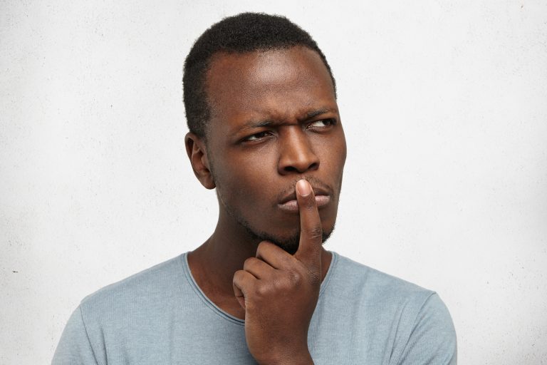 Close up studio portrait of handsome young black man having concentrated thoughtful expression, frowning, keeping finger on his lips as if trying to remember something or saying: And what if